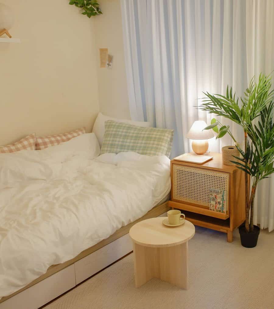 Small bedroom with bed, checkered pillows, nightstand, potted plant, and lamp; small round table with a green mug in foreground