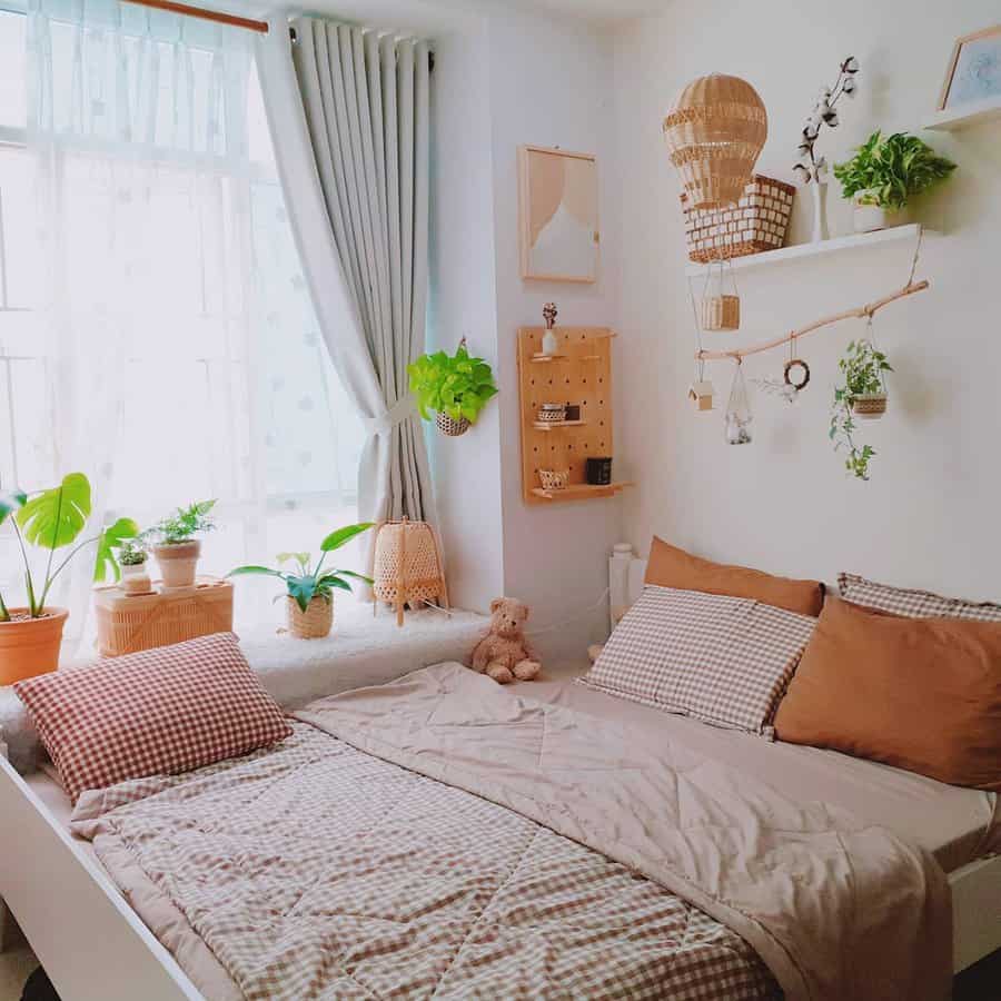 Cozy bedroom with a neatly made bed, warm-toned pillows, potted plants, a teddy bear, and soft natural light from a window