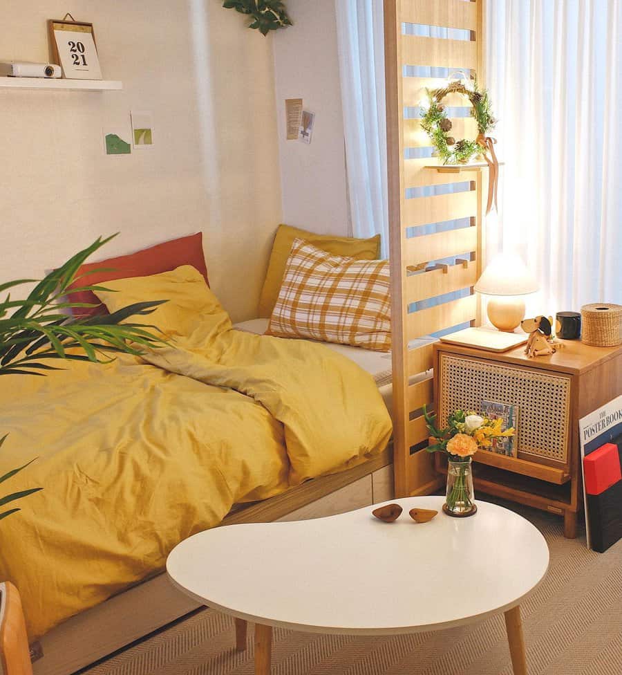 Small bedroom with a yellow bedspread, a wooden divider, a side table with a lamp, flowers, and a small white coffee table