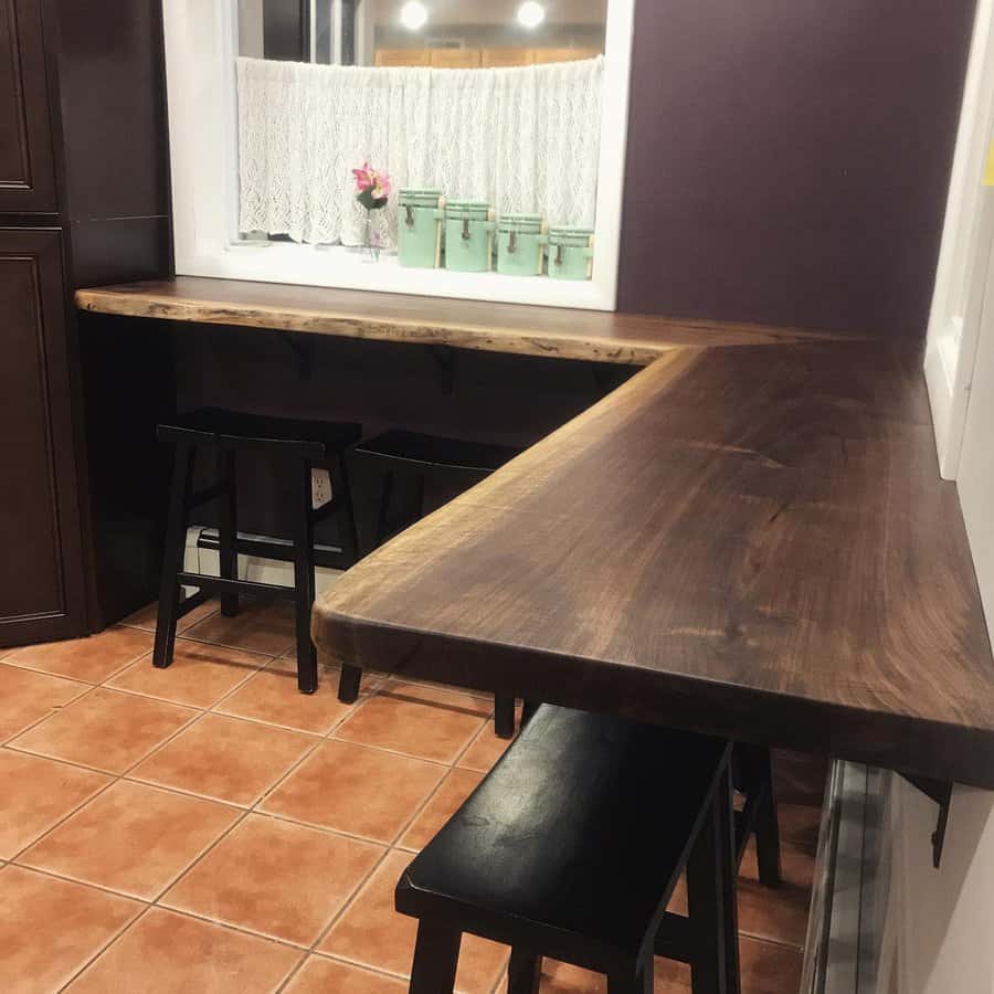 Wooden L-shaped breakfast bar with black stools on a tiled floor, near a window with lace curtains and green containers