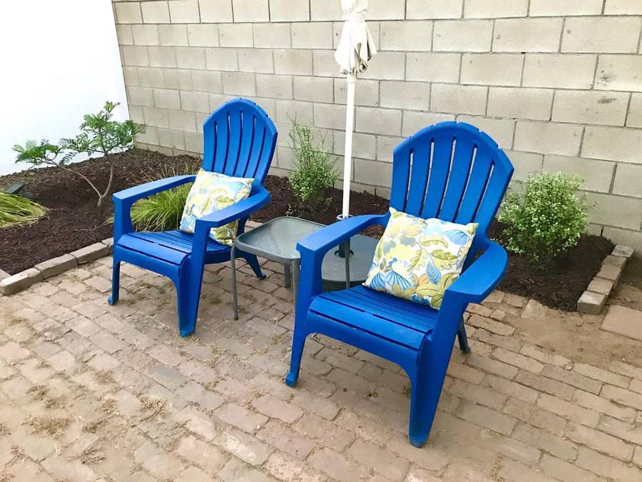 Two blue Adirondack chairs with floral cushions beside a small table with an umbrella, set on a patio with plants and brick flooring