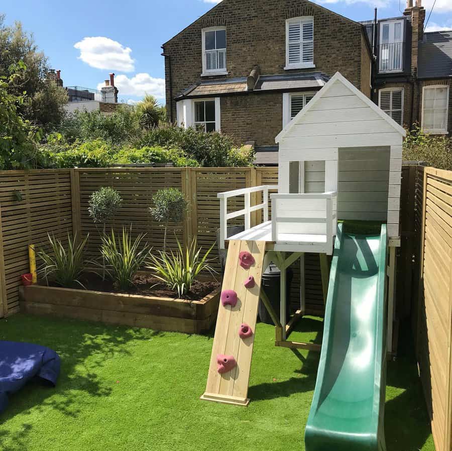 Small backyard with artificial grass, a white playhouse, a green slide, climbing wall, and a planter with shrubs