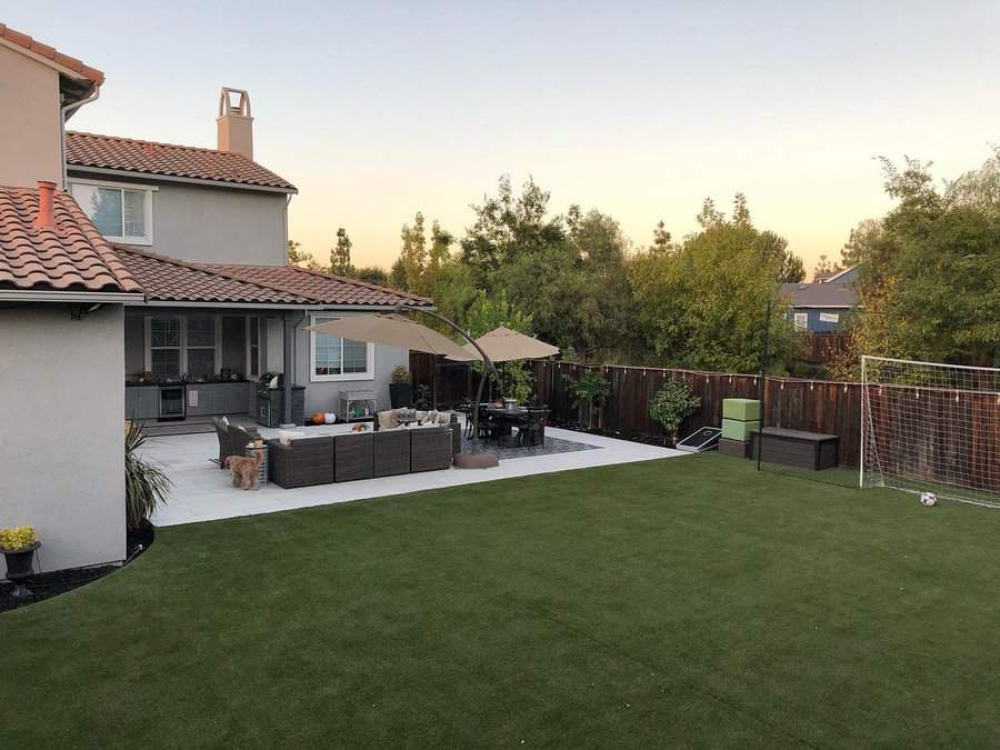 Backyard with artificial turf, patio seating, parasols, a soccer goal, and a wood fence; trees are in the background