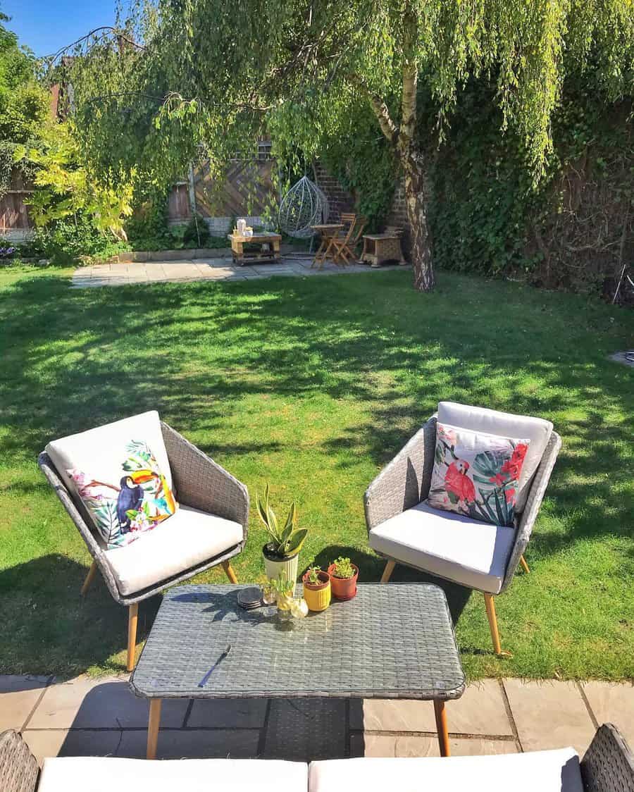 Outdoor garden with two cushioned chairs, a wicker table, and potted plants. Green lawn and trees in the background