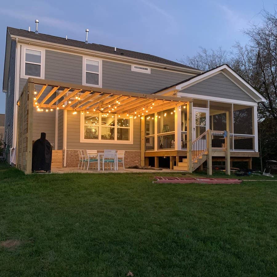 Two-story house with patio lights and seating area. Wooden pergola and string lights illuminate the backyard at dusk