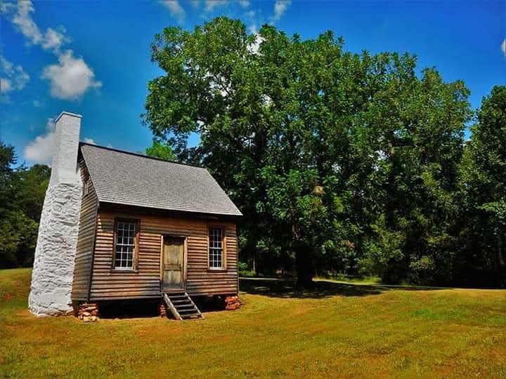 Historic wooden cabin with a white chimney surrounded by lush trees