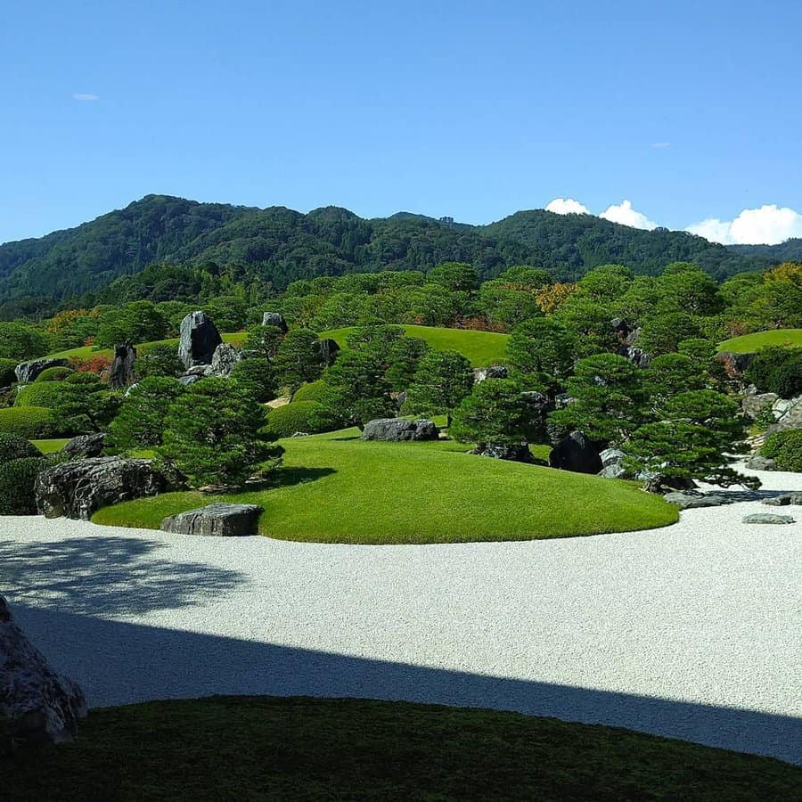 Zen garden with manicured shrubs, rocks, and gravel under a clear blue sky, surrounded by lush green mountains