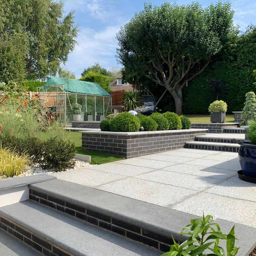 Modern low-maintenance backyard featuring raised planters with neatly trimmed shrubs, a tree centerpiece, and a greenhouse in the background, perfect for a structured yet inviting landscape