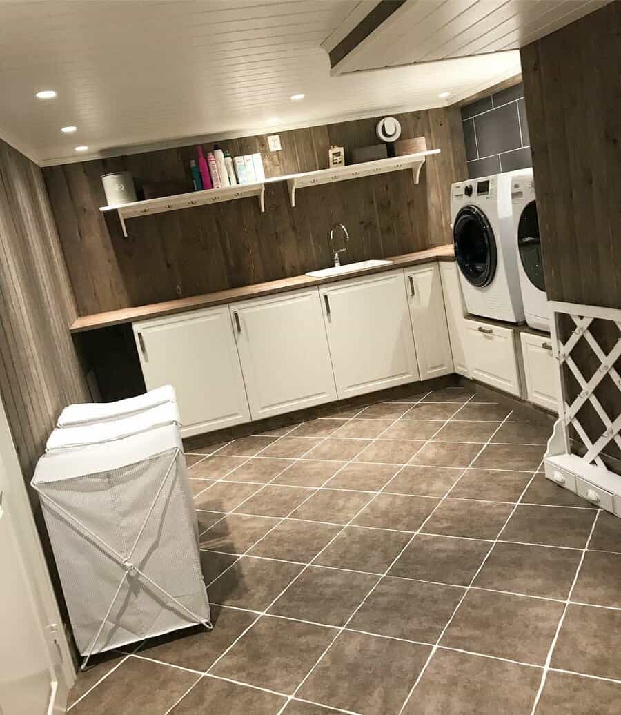 Rustic basement laundry room with wood panel walls, white cabinetry, a built-in sink, and ample storage for a functional and stylish space.