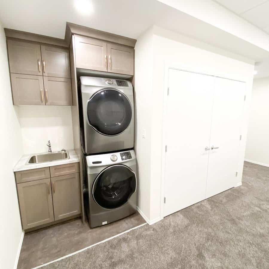 Compact basement laundry nook with stacked stainless steel washer and dryer, built-in cabinetry, and a small sink for a sleek and efficient setup.