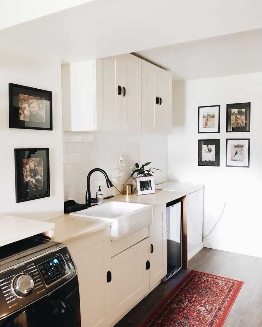 Charming basement laundry room with a farmhouse sink, white cabinetry, black hardware, and a cozy touch with framed family photos and a rug.