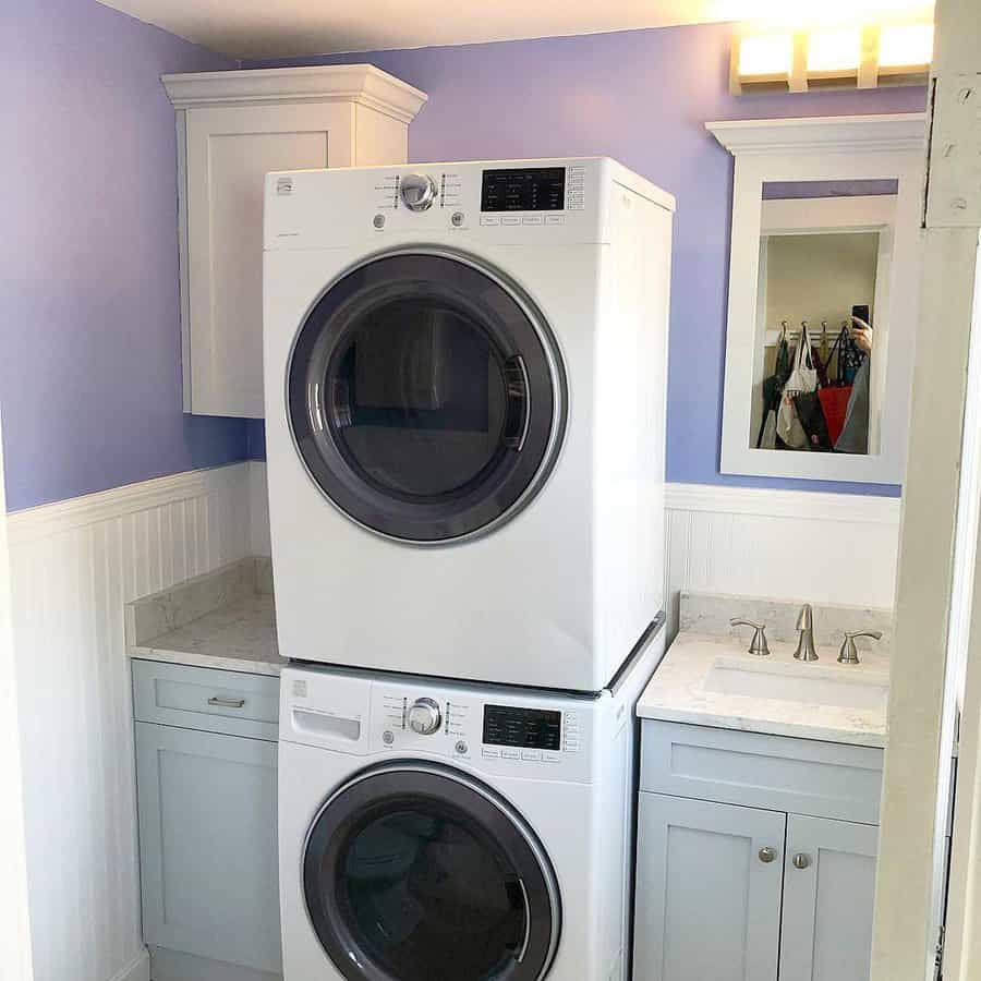 Stacked washer and dryer in a small laundry room with light blue walls, white cabinets, and a sink with a mirror above