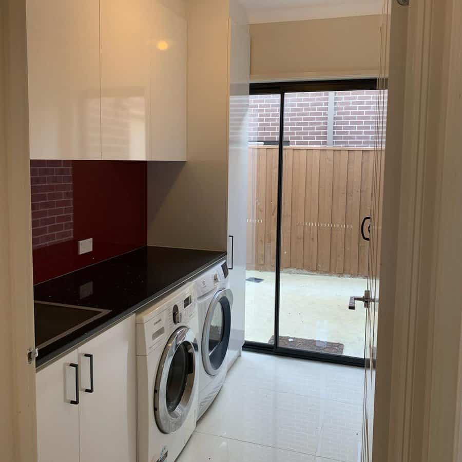 Laundry room with a washer and dryer under a black countertop, red backsplash, white cabinets, and a glass door leading outside