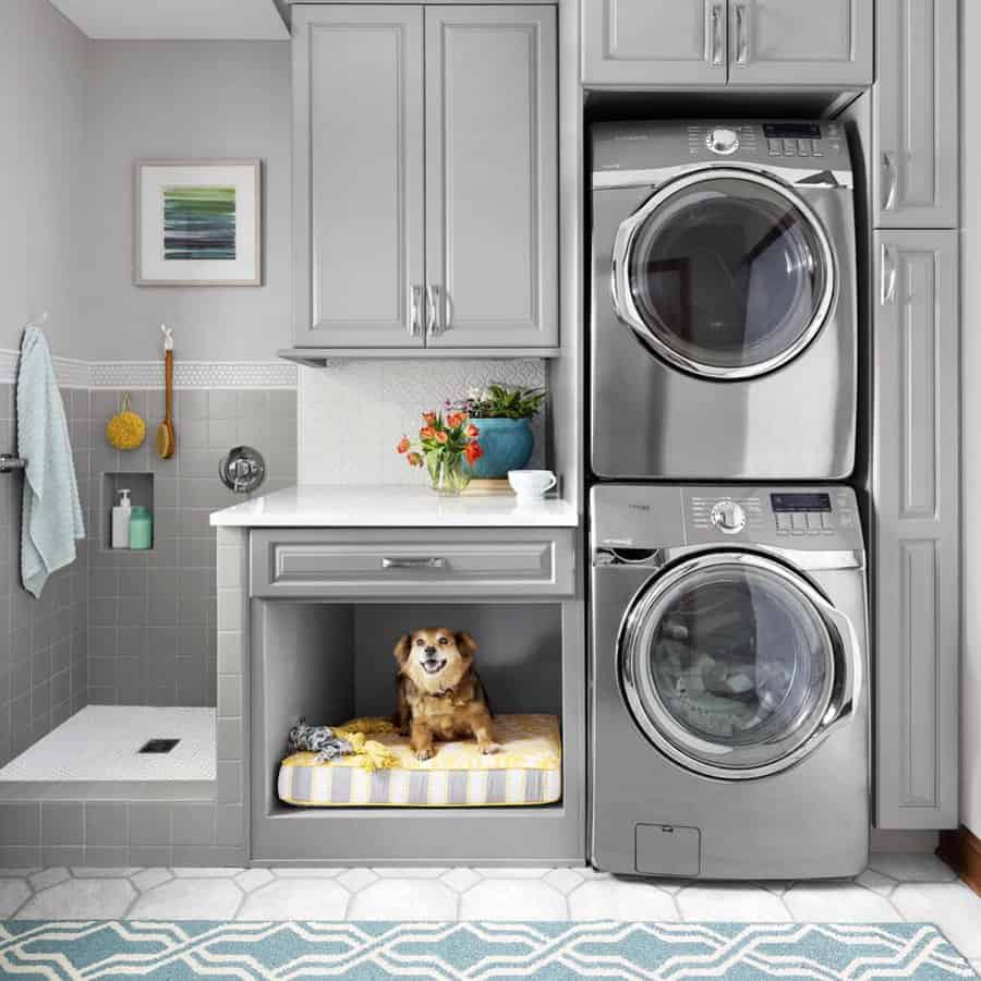 Laundry room with stacked washer and dryer, gray cabinets, a dog on a bed in a kennel area, and decor on a countertop