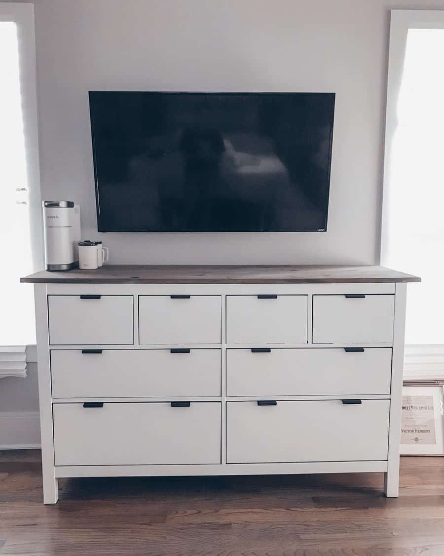 White dresser with nine drawers beneath a wall-mounted flat-screen TV; a mug and tumbler are on the dresser in a light-filled room