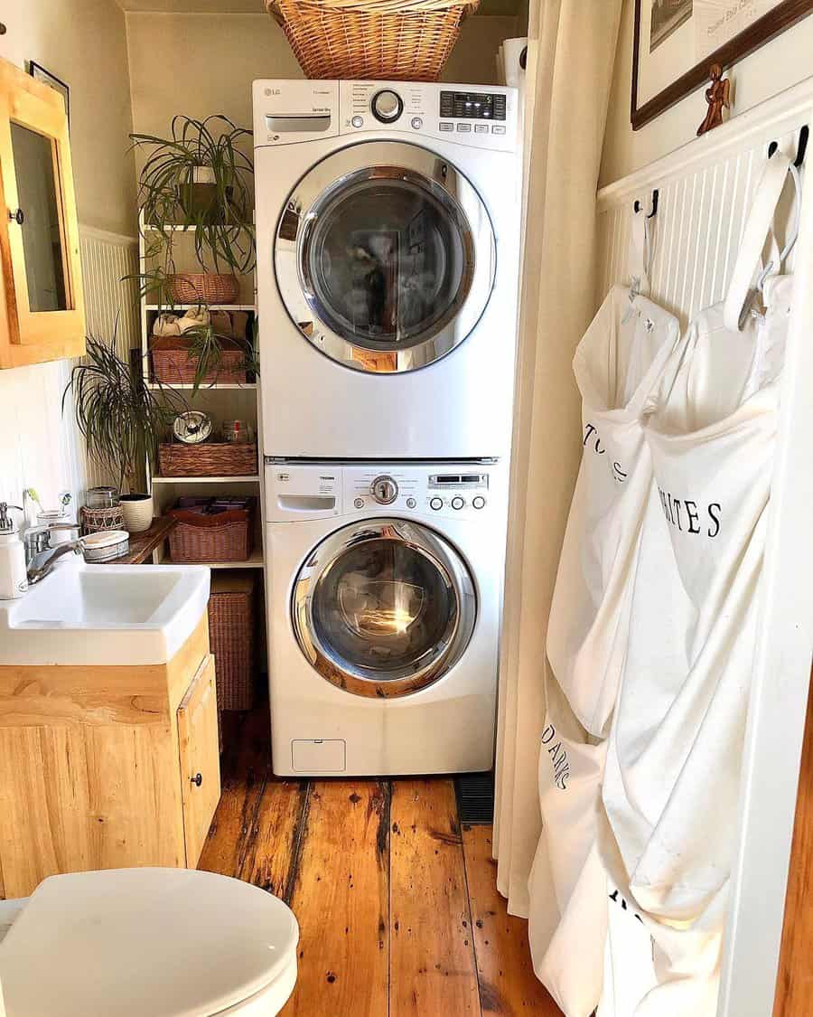 A compact laundry room with stacked washer and dryer, plants, and a wooden cabinet, with laundry bags hanging on a nearby wall