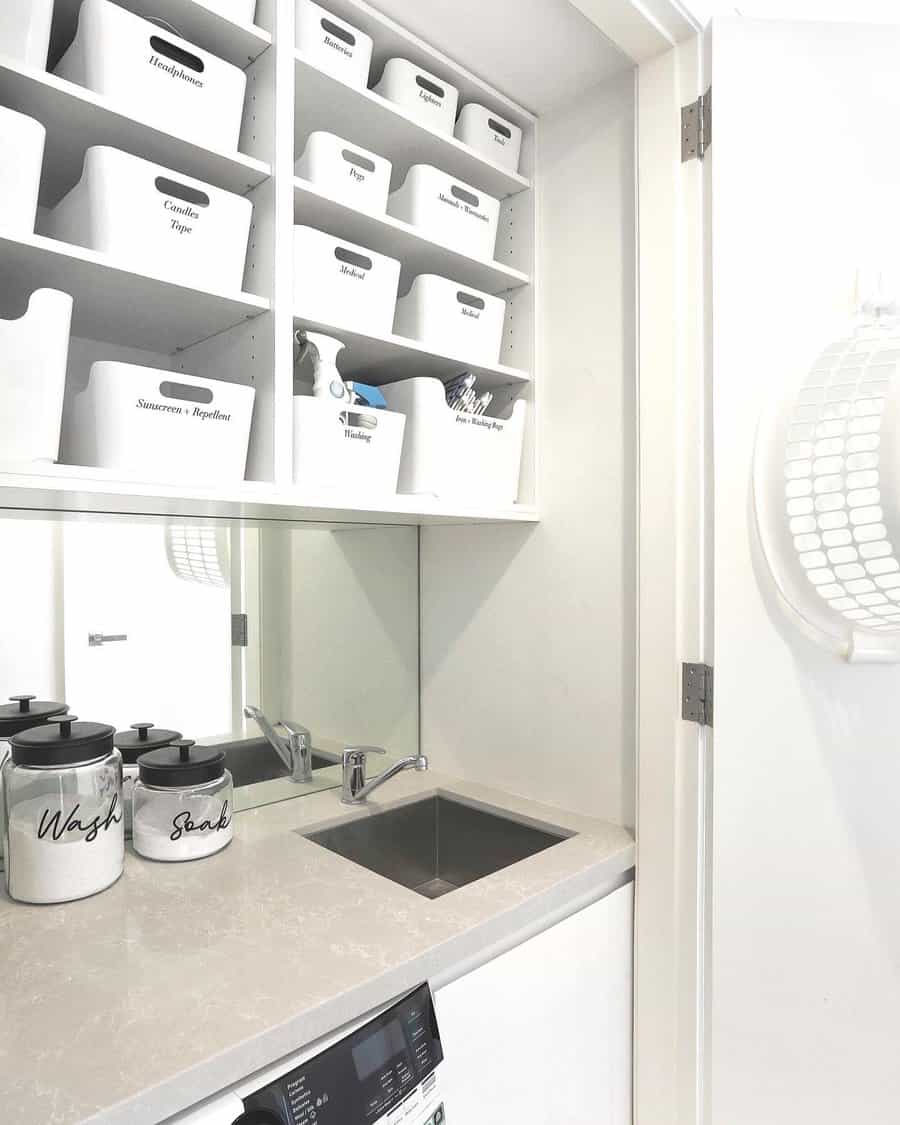 Compact laundry room with labeled white storage bins on shelves, a small sink, and washing machine; soap containers are on the counter