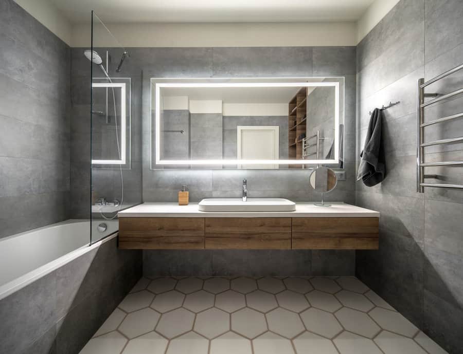 Modern bathroom with a large mirror, wooden vanity, bathtub, hexagonal floor tiles, and gray walls