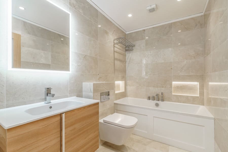 Modern bathroom with beige tiles, illuminated mirror, wooden vanity, wall-mounted toilet, and bathtub