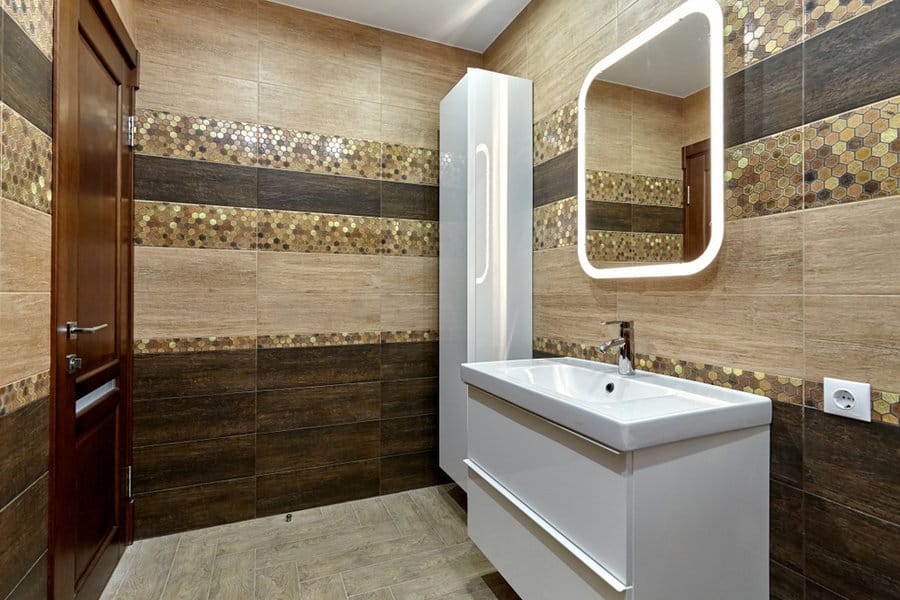 Modern bathroom with brown tile walls, illuminated mirror, white sink, and a wooden door; hexagonal tile accents add texture