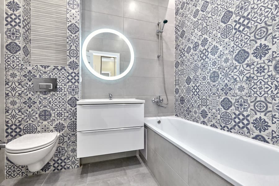 Bathroom with patterned tiles, a round mirror above a white sink, wall-mounted toilet, and a bathtub with a handheld showerhead