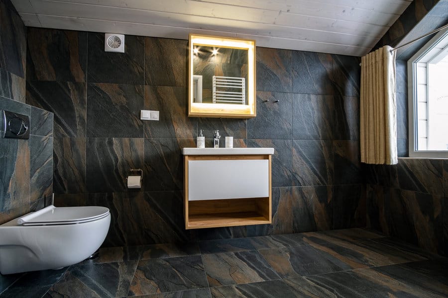 Modern bathroom with dark stone walls and floor, wall-mounted toilet, wooden vanity, illuminated mirror, and window with curtains