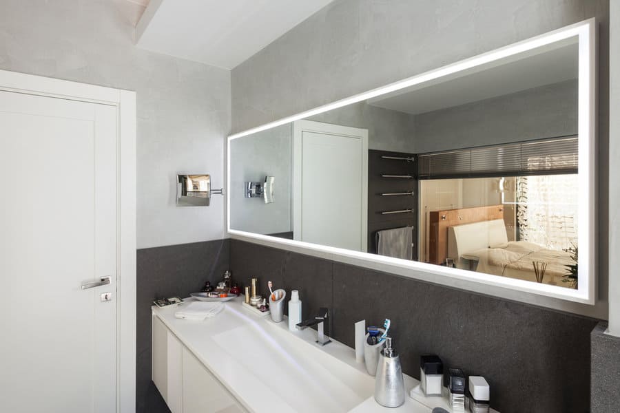 Modern bathroom with a large illuminated mirror, white sink, and various toiletries on the counter, light gray walls and a towel rack