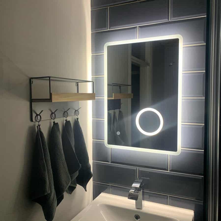 Bathroom with illuminated mirror, dark tiles, white sink, modern faucet, and shelf with hooks holding three dark towels