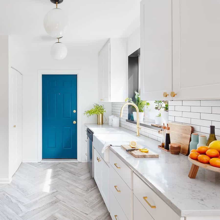 Bright kitchen with white cabinets and marble countertops, a blue door, fruit bowl, and plants add color, herringbone floor pattern