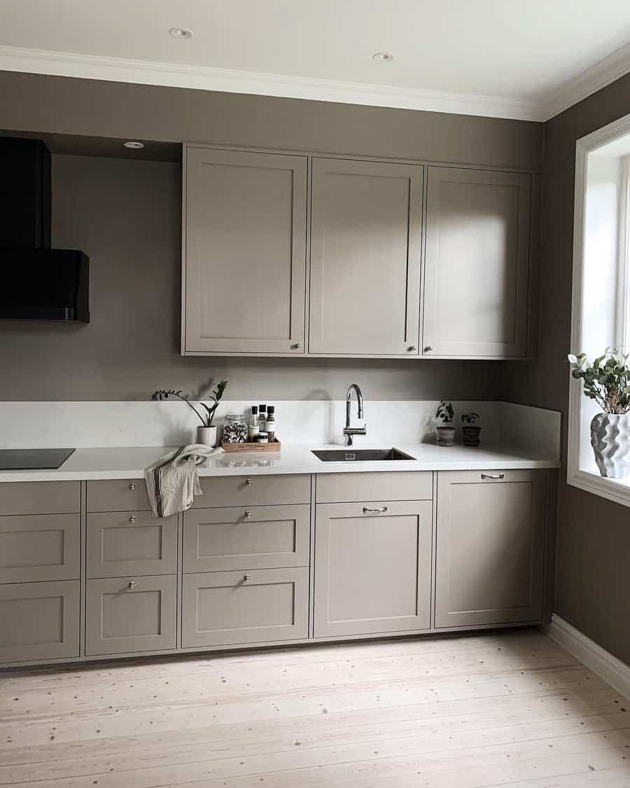 Minimalist kitchen with taupe cabinets, white countertops, and wood flooring featuring a small sink and vase on the counter, complemented by natural light from a window