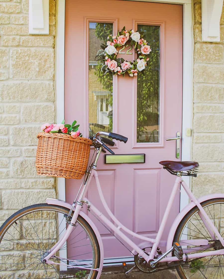 Pink bicycle with a wicker basket filled with flowers, leaning against a pink door adorned with a floral wreath; stone wall backdrop