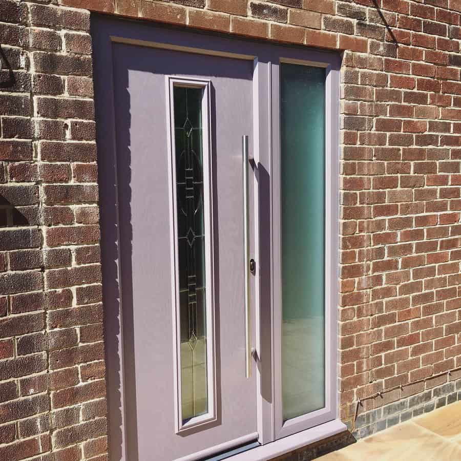 A purple door with a patterned glass insert and side light is set in a red brick wall, with sunlight casting shadows