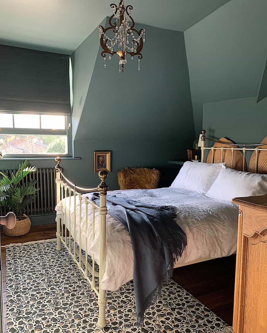 Cozy bedroom with a vintage bed, green walls, chandelier, and window; a plant and patterned rug add warmth to the decor