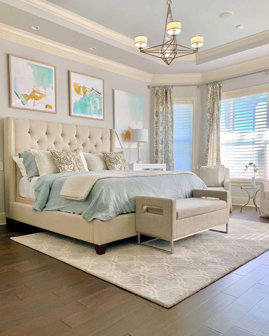 Spacious master bedroom with a large bed, tufted headboard, abstract art, chandelier, cushioned bench, and elegant curtains