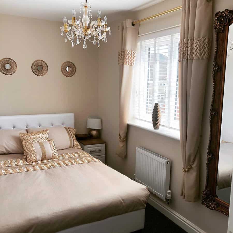 Elegant bedroom with a beige color scheme features a bed, ornate mirror, chandelier, decorative wall art, and large window with curtains