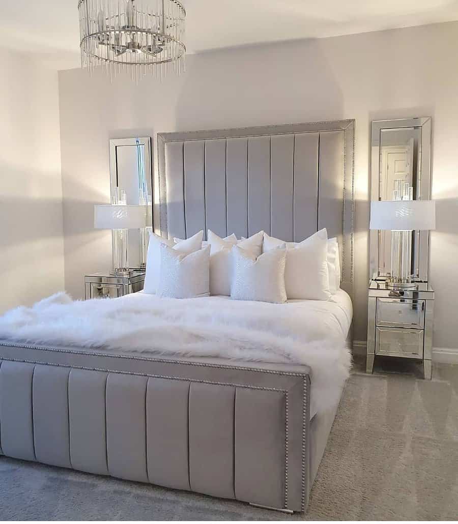 Elegant bedroom with a tall, upholstered bed, fluffy white bedding, mirrored side tables, modern lamps, and a chandelier