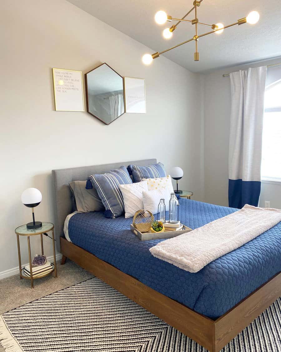 Modern bedroom with wooden bed, blue quilt, and pillows; round tables with lamps, geometric wall art, and a gold chandelier overhead