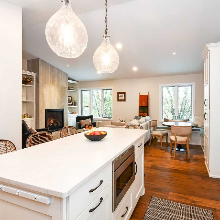 Modern kitchen and living area with white cabinets, wooden floor, large island, pendant lights, and a cozy fireplace in the background