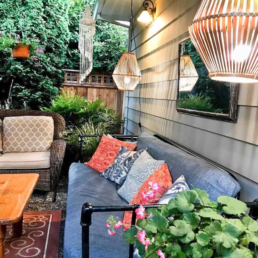 Cozy patio with a wicker chair and a blue sofa adorned with colorful pillows, surrounded by greenery and warm pendant lighting