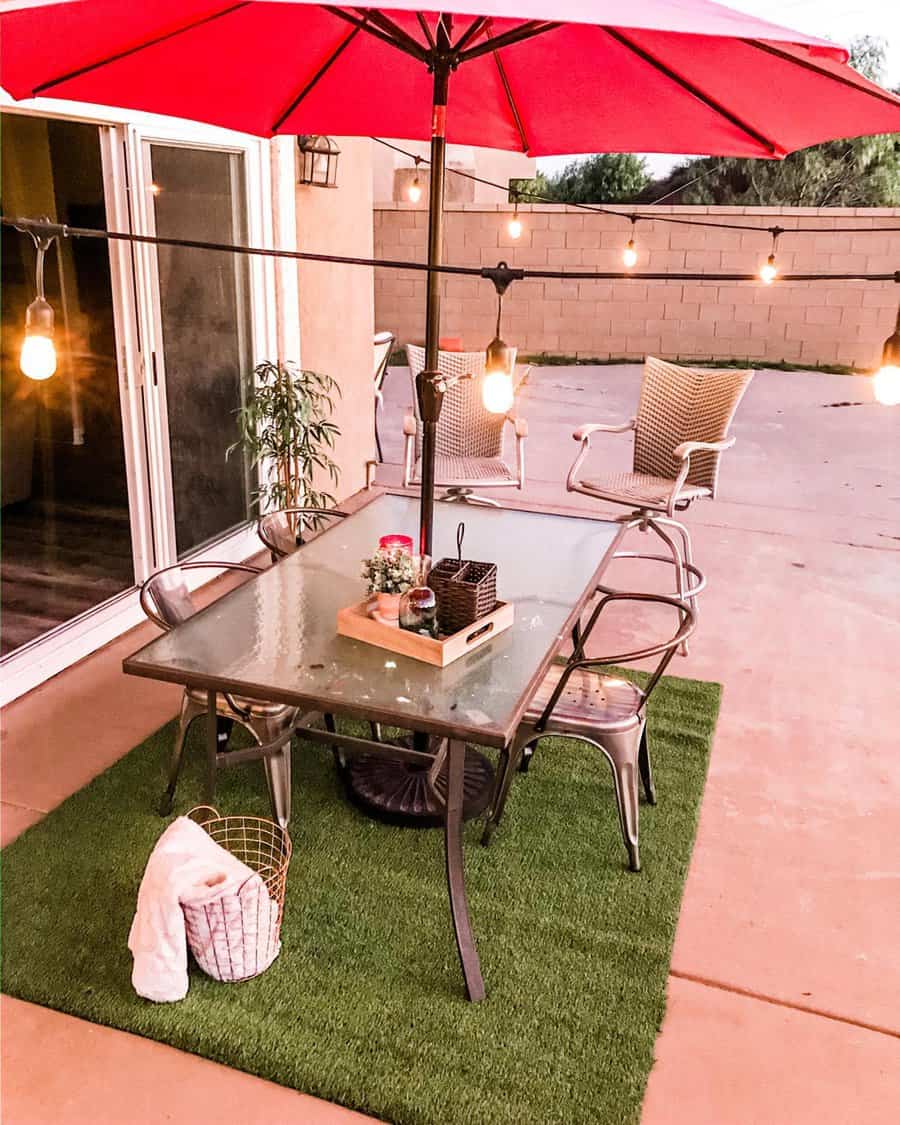 Patio with a glass table, red umbrella, metal chairs, string lights, and a small rug. A basket with a towel is on the ground