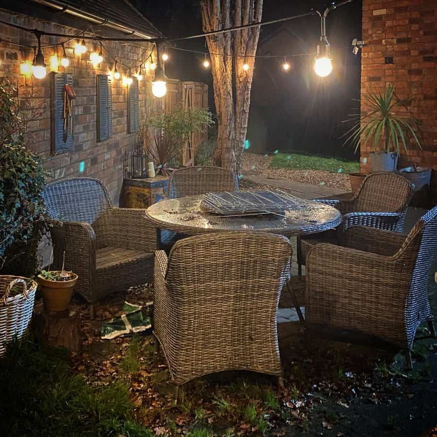 Outdoor patio with wicker chairs around a round table, string lights above, and a brick wall on the side at night