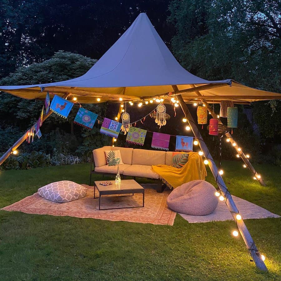 A cozy outdoor tent setup with string lights, colorful decor, a sofa, and a table on a rug in a grassy area during evening time