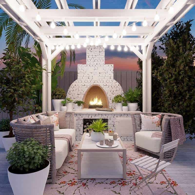 Outdoor patio with white pergola, string lights, wicker furniture, and a white brick fireplace at sunset, lush plants surround