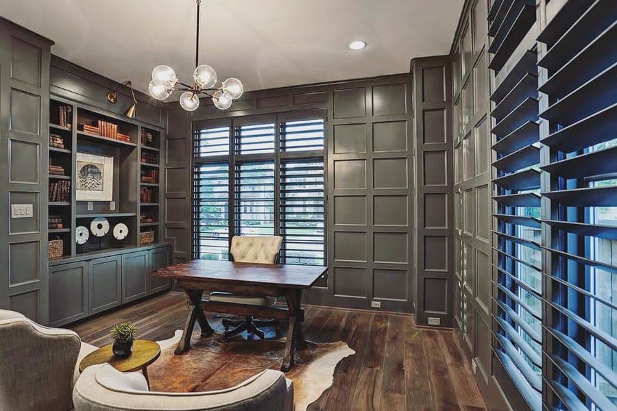 A modern office with a wooden desk, beige chair, built-in shelves, geometric light fixture, and large windows with dark shutters