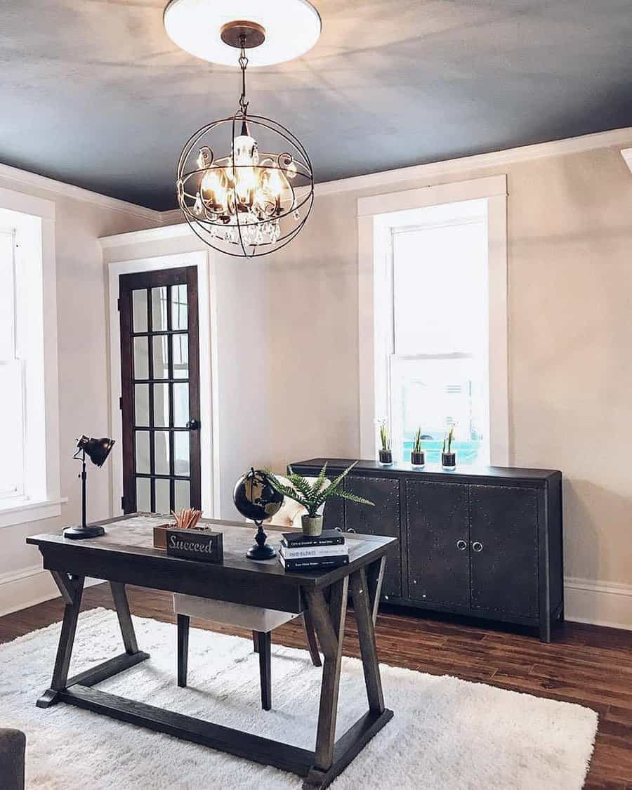 A stylish home office with a dark wood desk, globe, and books, complemented by a modern chandelier, cabinet, and large windows on a white rug