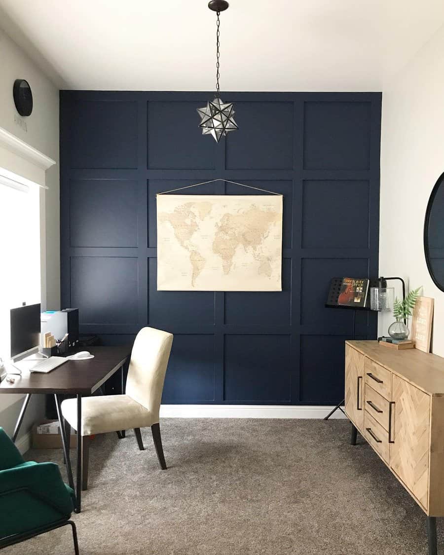 Minimalist home office with a dark-paneled accent wall, world map, wooden desk, beige chair, pendant light, and sideboard