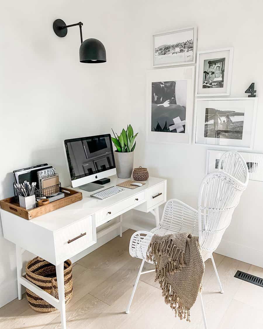 Minimalist home office with a white desk, wicker chair, computer, potted plant, wall art, and a black wall lamp in a neutral decor theme