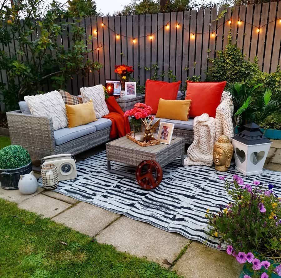 Outdoor seating area with gray sofas, red and orange cushions, a table, lanterns, potted plants, and string lights above on a patio