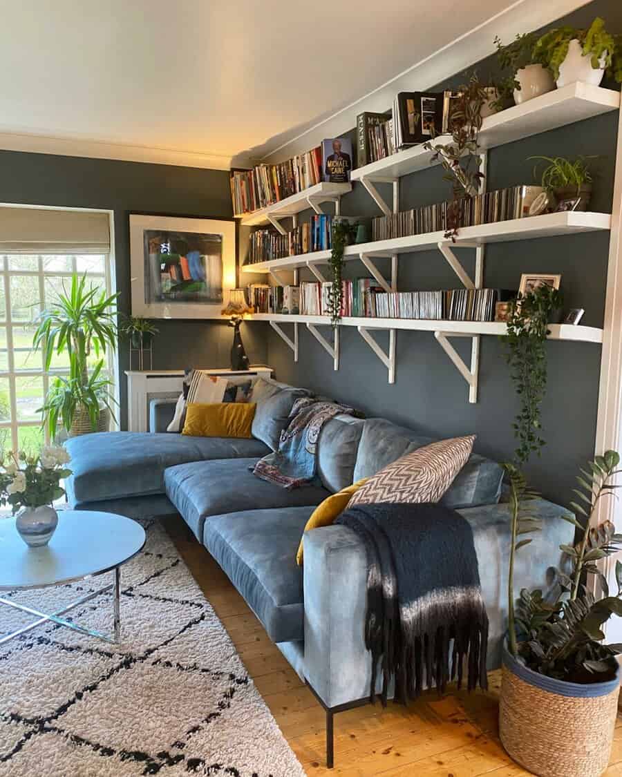 Cozy living room with floating white bookshelves filled with books, CDs, and plants, a plush blue sectional sofa, and warm decor accents.