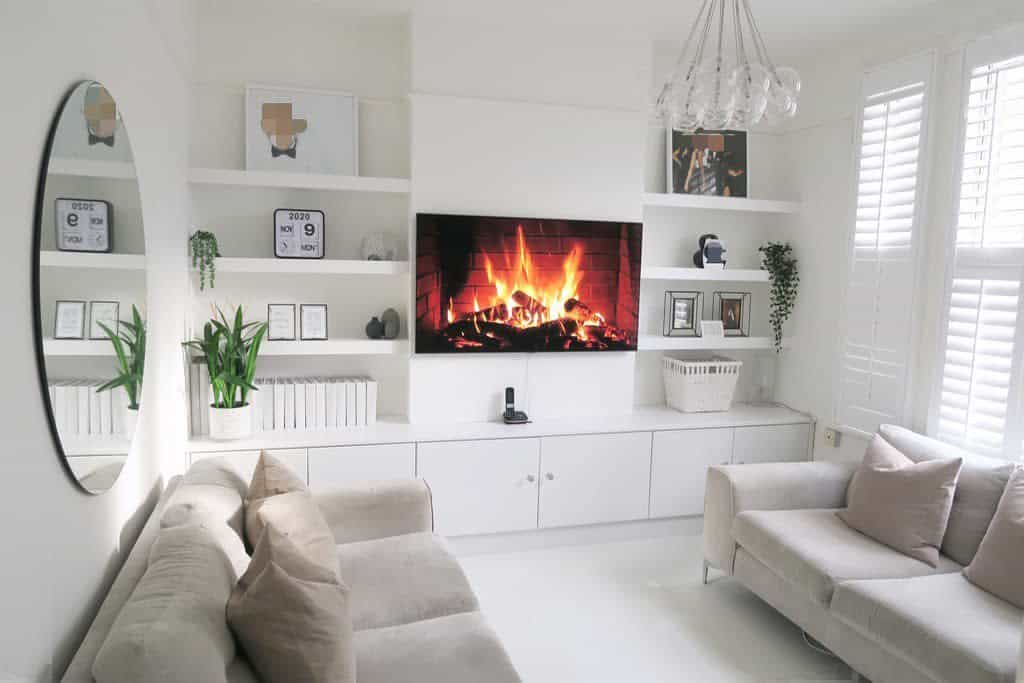 Minimalist white living room with built-in floating shelves, a wall-mounted fireplace TV, soft neutral sofas, and modern decor